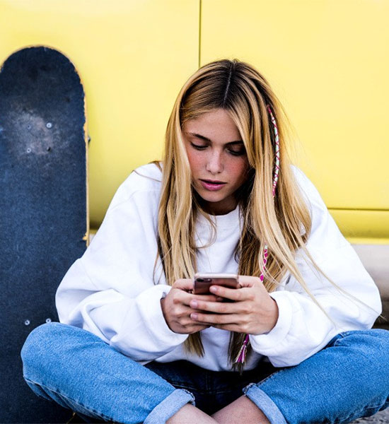 Eine junge Frau sitzt vor einer gelben Wand auf dem Boden neben einem Skateboard und schaut auf ihr Smartphone.