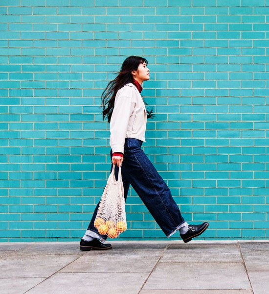 Eine Frau geht an einer türkisfarbenen Wand entlang, sie trägt eine Tragetasche mit Orangen.
