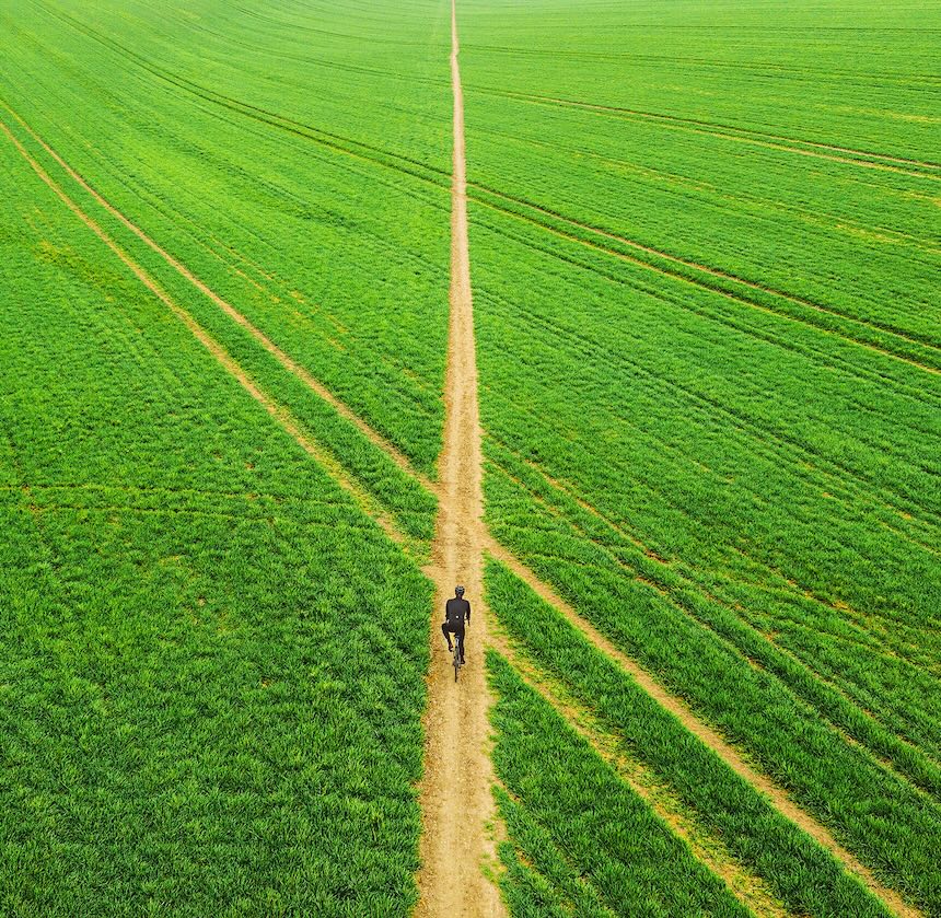 Person walking along a narrow dirt path through a vast green field, viewed from above.