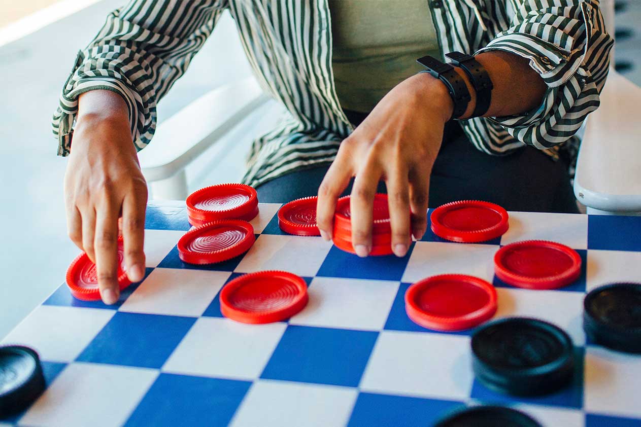 Une personne assise à une table jouant aux dames, les mains positionnées au-dessus du plateau dans un moment de concentration et de stratégie.