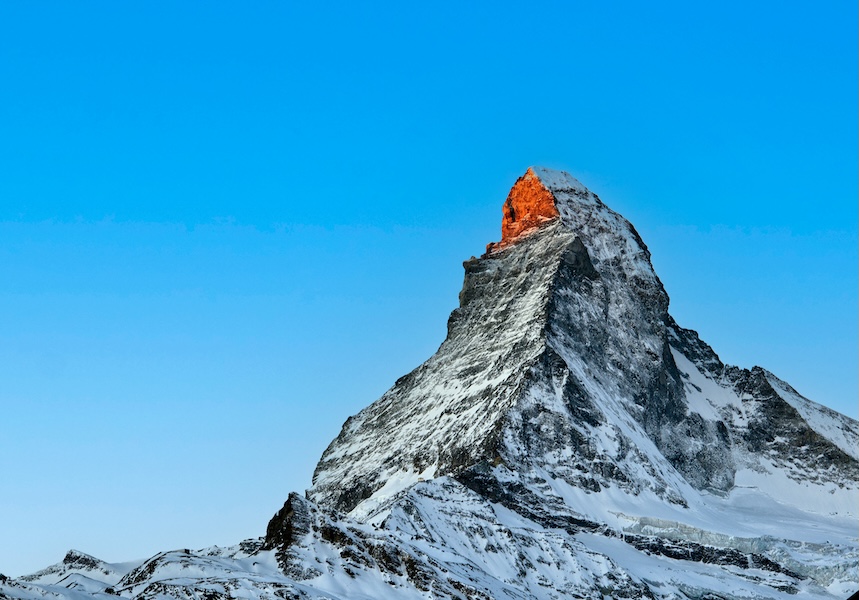 Schneebedeckter Berggipfel mit orangefarbenem Sonnenlicht vor blauem Himmel