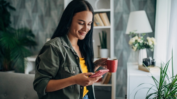 Une personne se tient dans un intérieur moderne, tenant un smartphone et une tasse, dans une scène du quotidien calme et contemporaine.