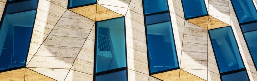 Close-up of a modern building façade featuring angular beige stone panels and tall blue-tinted windows arranged in an irregular geometric pattern. The contrasting shapes and reflective glass create a contemporary architectural design.