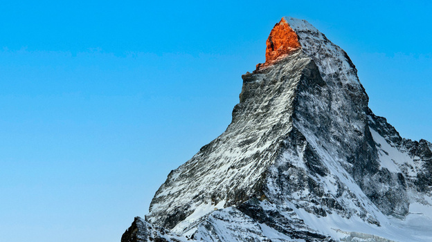Snow-covered mountain peak illuminated by warm sunlight against a clear blue sky.