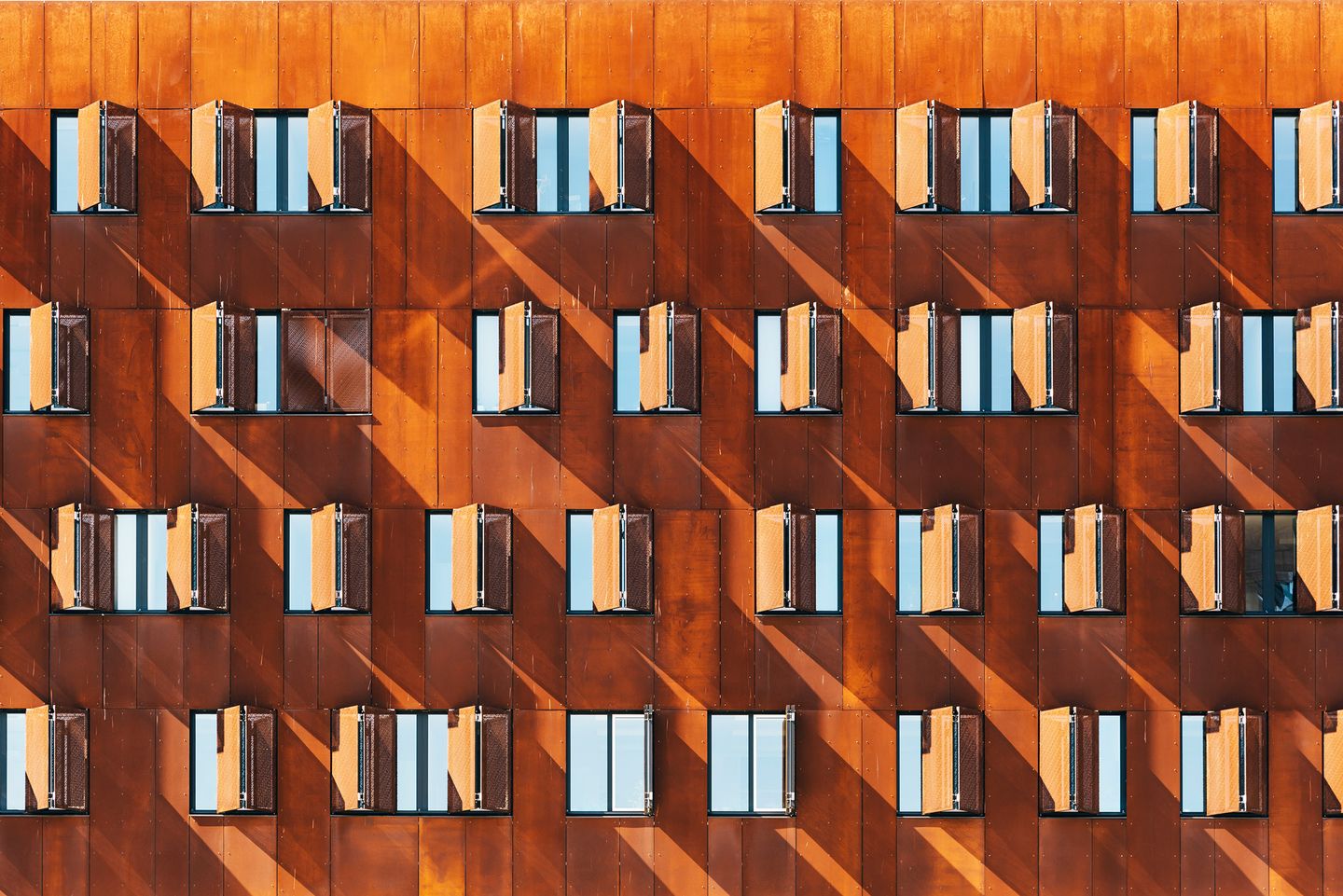 Rust-colored building facade with rows of shuttered windows in sunlight.