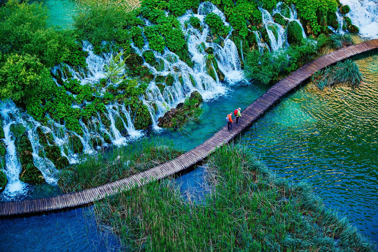 Hikers on a wooden boardwalk over clear water with waterfalls and greenery.