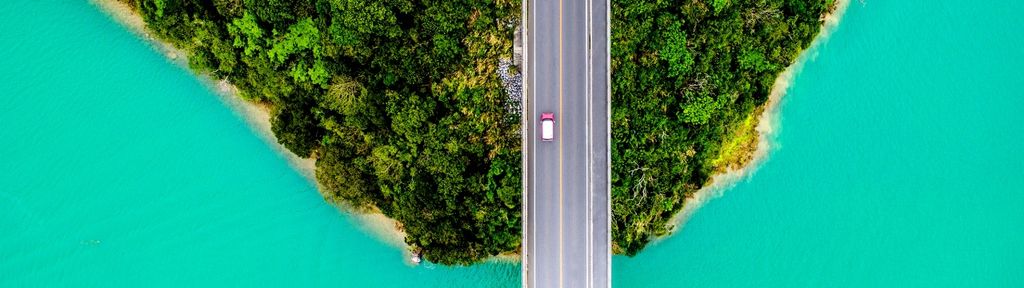 Aerial view of a vehicle driving across a bridge spanning turquoise water, bordered by lush green vegetation on both sides.