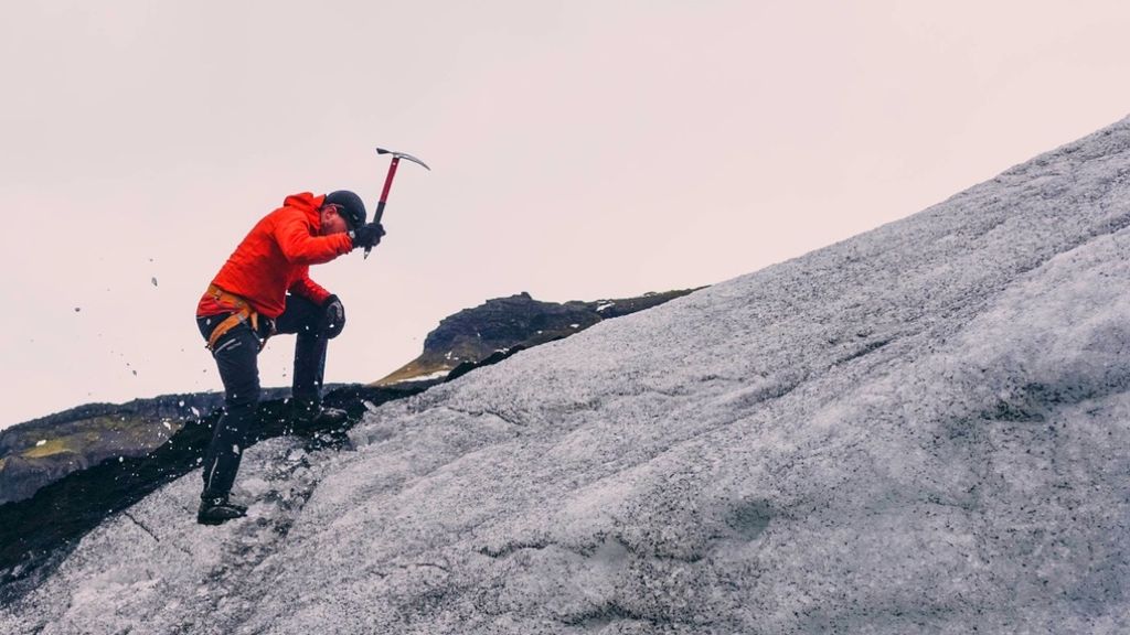 A person in an orange jacket and climbing gear uses an ice axe to climb a snowy slope.