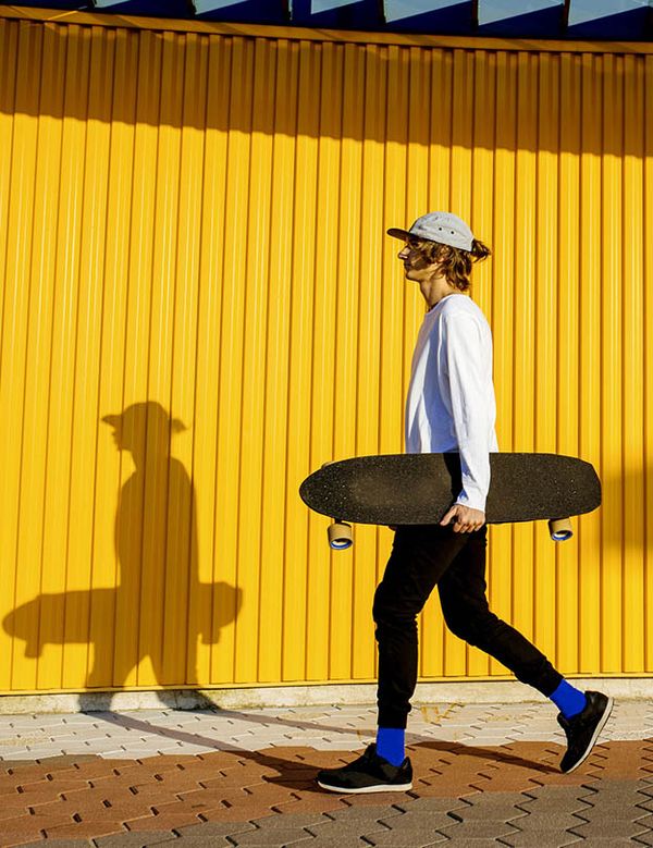 Mladý muž se skateboardem v podpaží kráčí podél žluté zdi.