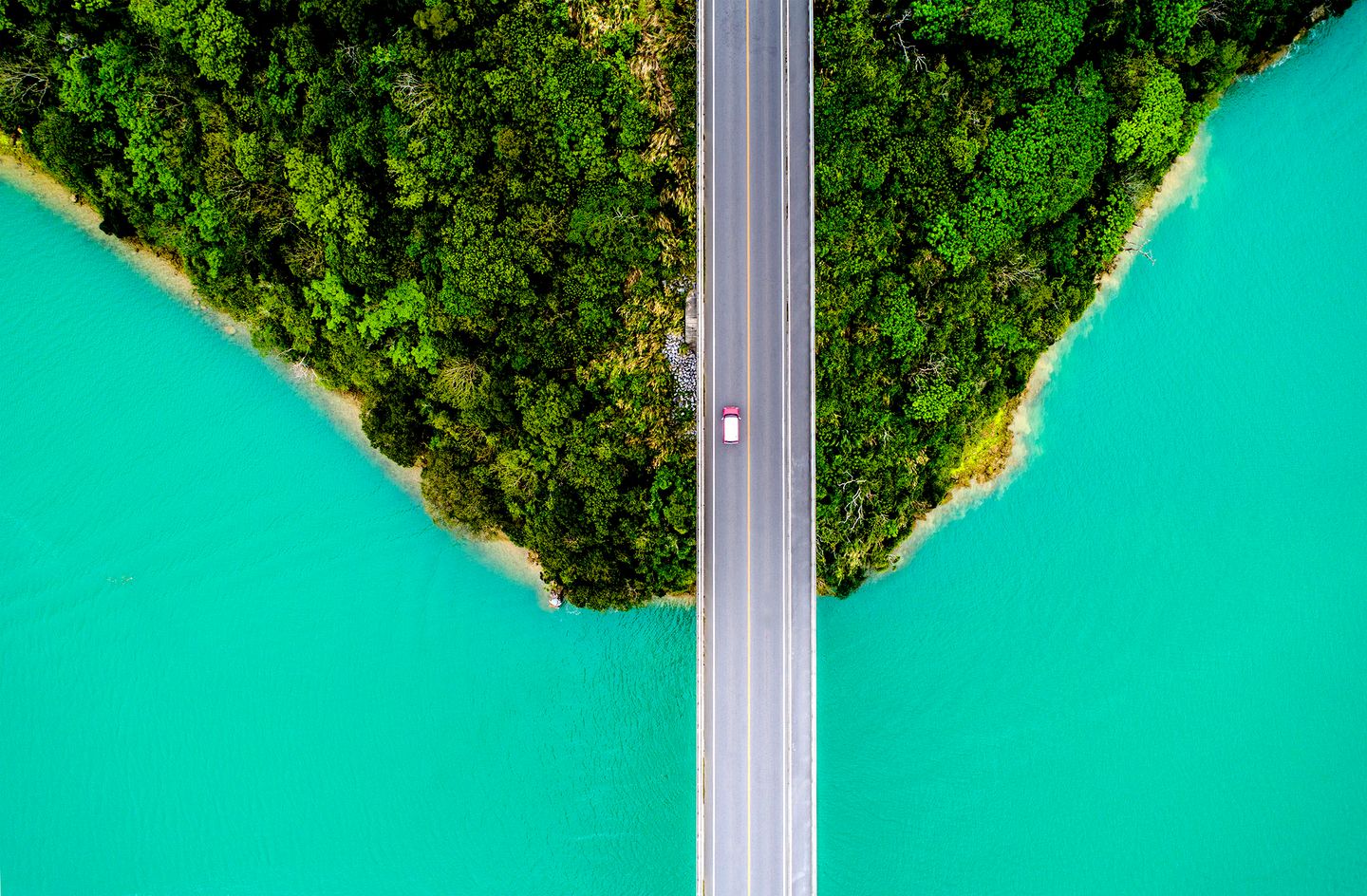 Aerial view of a bridge with a single car crossing between dense green forest and bright turquoise water..