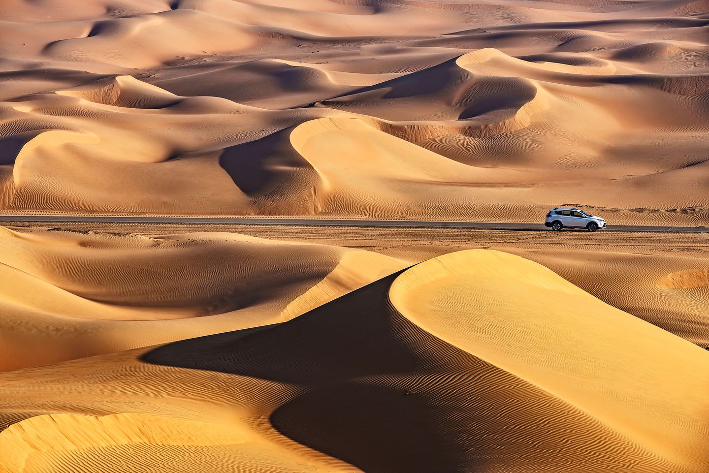 Silver car driving along a road surrounded by tall golden sand dunes in a vast desert landscape.