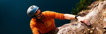 A person wearing a grey climbing helmet and dark sunglasses, looking up while rock climbing