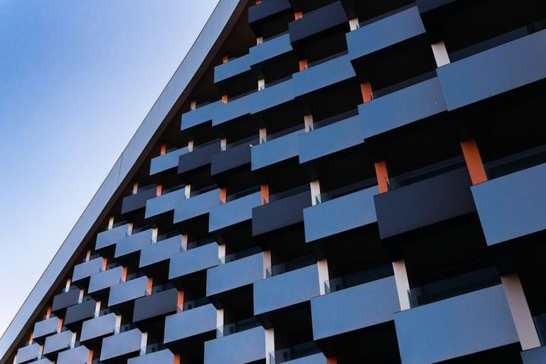 A building facade with a series of staggered rectangular balconies in dark blue and gray tones, creating a geometric pattern. The structure is angled upward toward a clear blue sky.