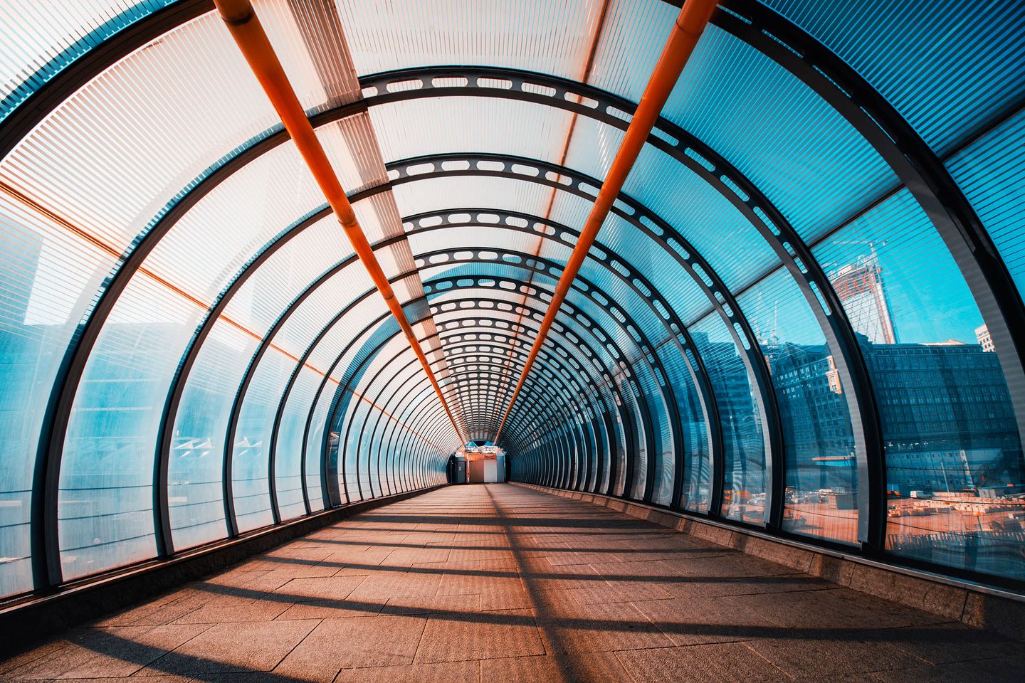 Interior of a curved glass walkway tunnel with metal supports, sunlight casting long shadows on the floor.