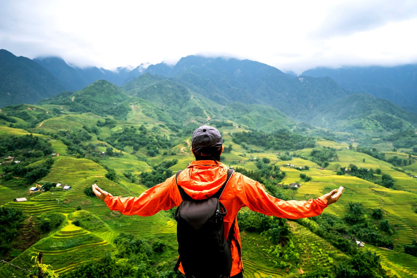 Person in an orange jacket and backpack standing with arms open, overlooking green terraced hills and distant misty mountains