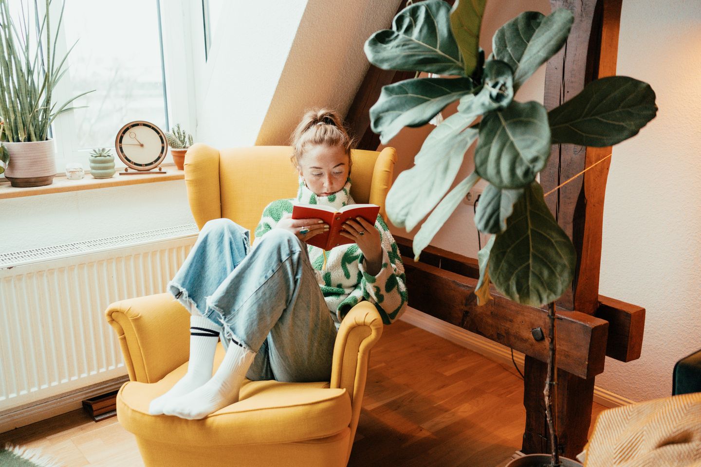 A woman in a green and white sweater and jeans sits in a yellow armchair reading a book near a window with potted plants.