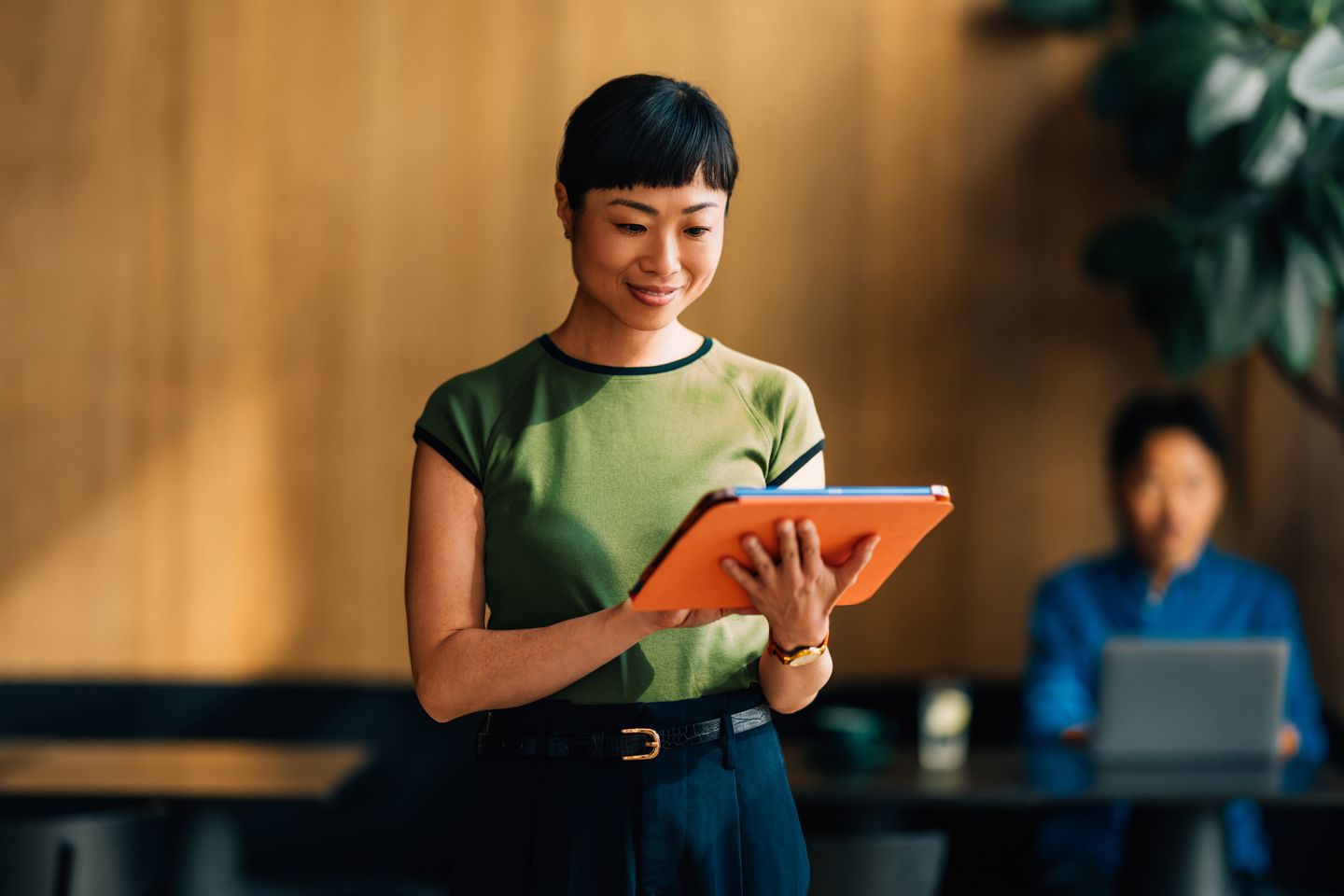 A woman standing indoors and using a tablet, with another person working in the background