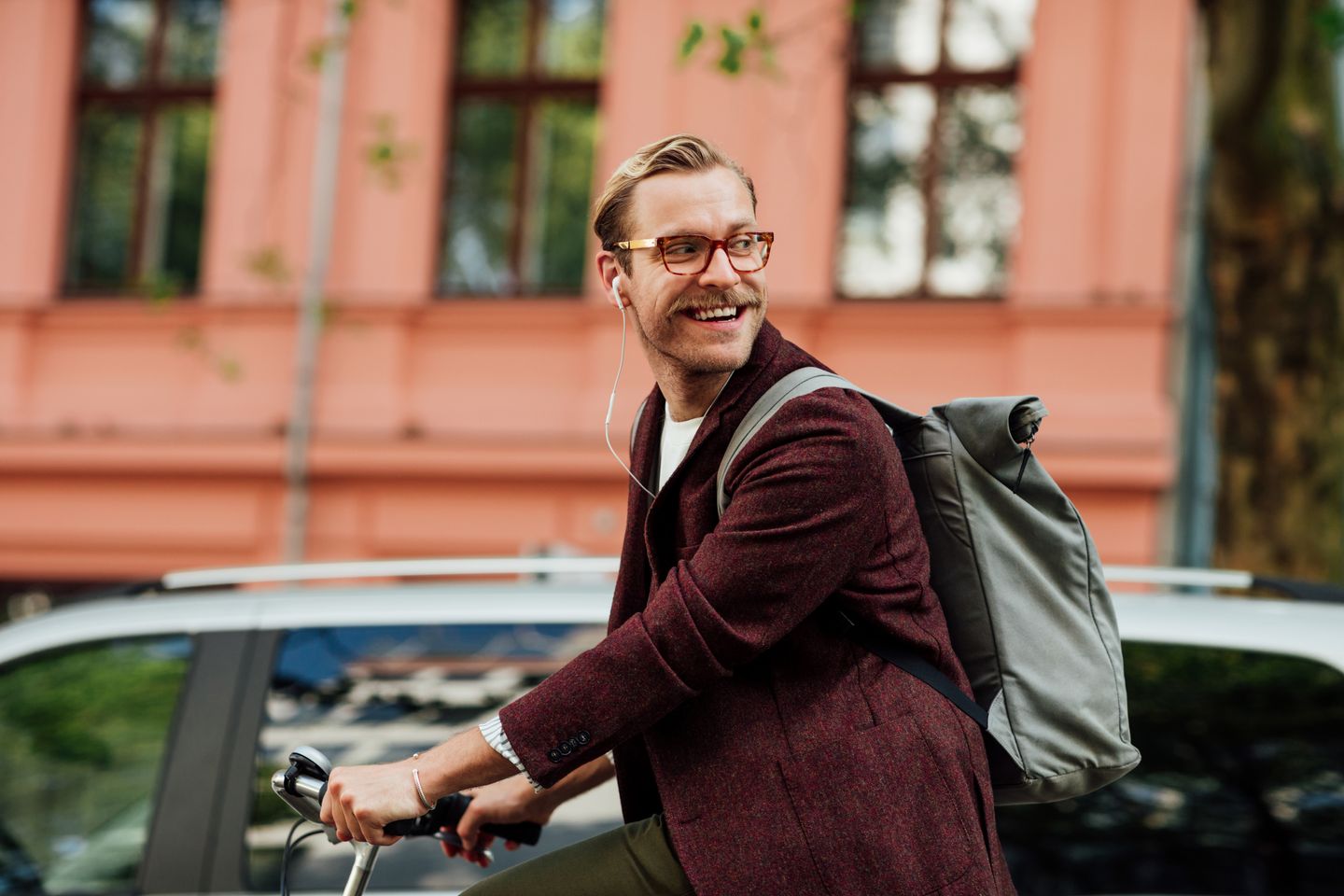 A man wearing glasses, earphones, and a dark coat rides a bicycle with a backpack in front of a building with large windows and greenery.